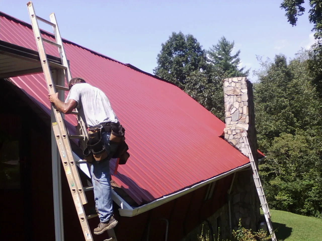 Roofer on a ladder beside a red metal roof and stone chimney — Soffit & Fascia Repair by Roofing Brothers in Old Orchard Beach